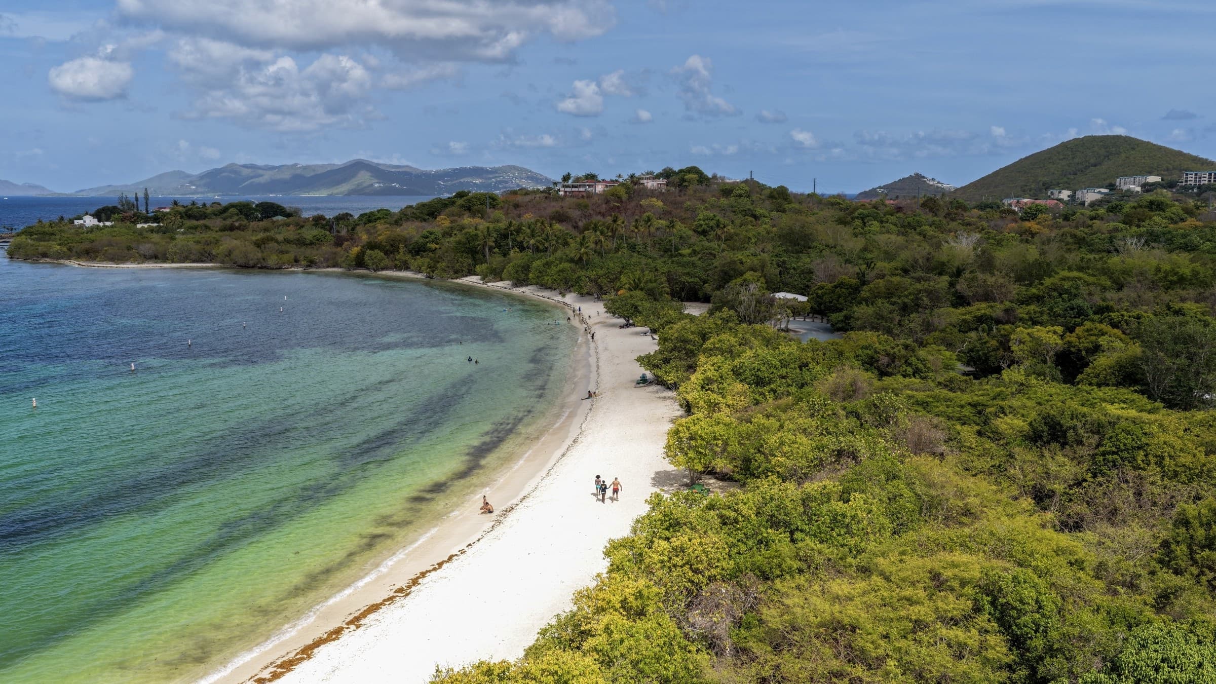 Aerial view along Lindquist Beach with shallow water, a white shoreline, and green hills on St. Thomas.
