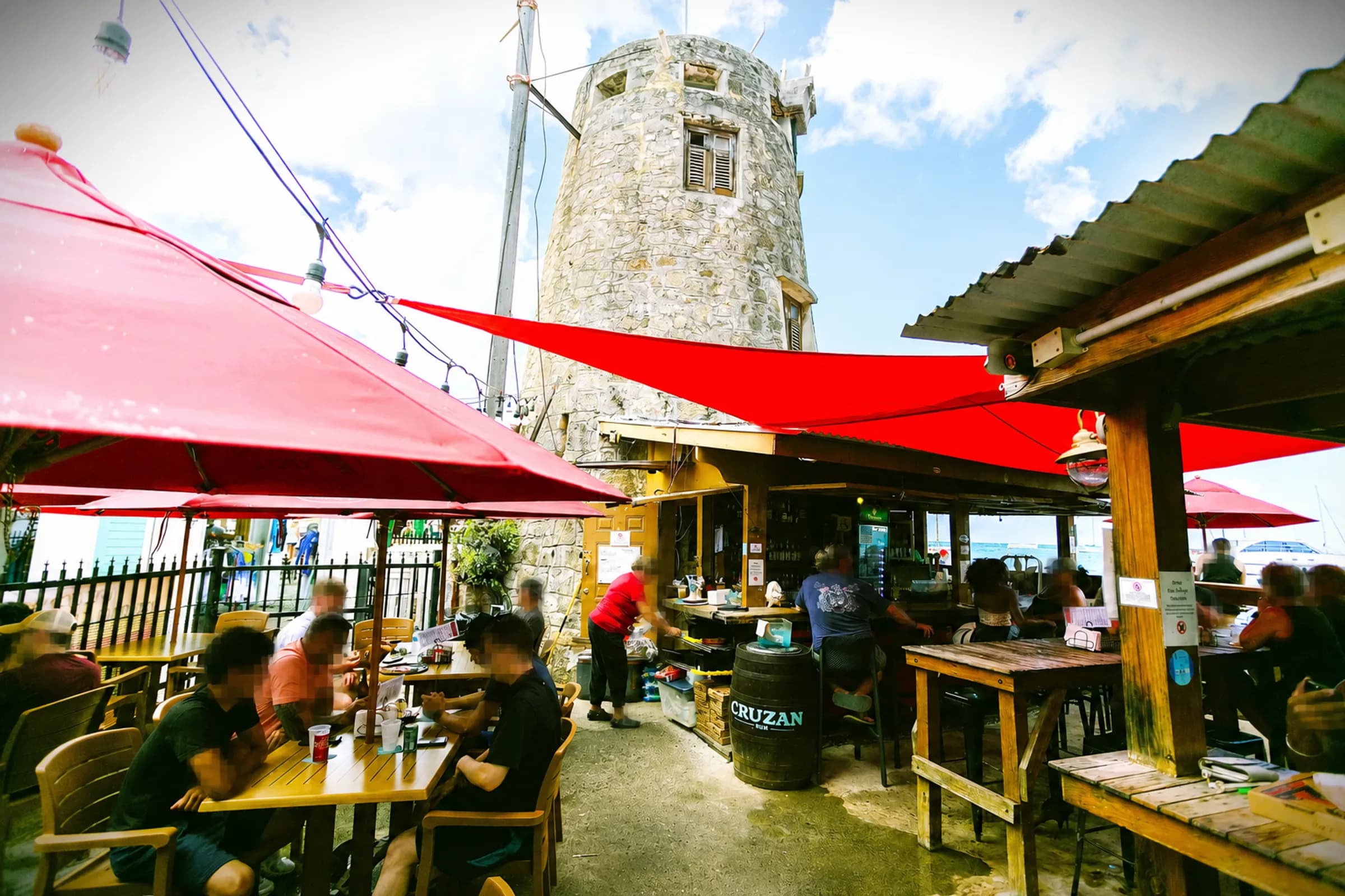 Open-air dining beneath red shade sails beside The Mill's historic tower.